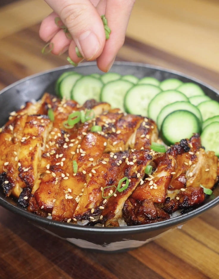 Fresh scallions being sprinkled over sliced Korean BBQ chicken and rice bowl with cucumbers and sesame seeds.