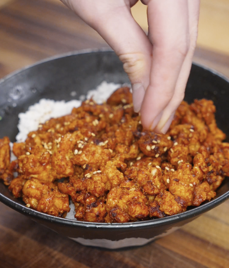 Hand sprinkling sesame seeds over Spicy Peanut Chicken served on rice, finishing the dish with texture and flavor.