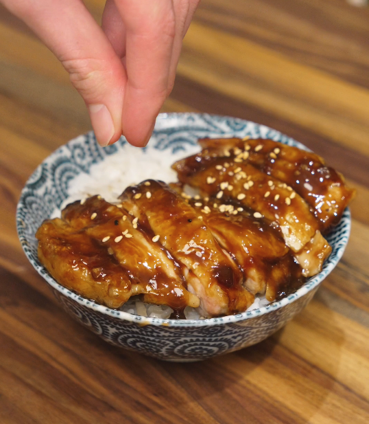 Sesame seeds being sprinkled over sliced teriyaki chicken served on rice, finishing the dish before serving.