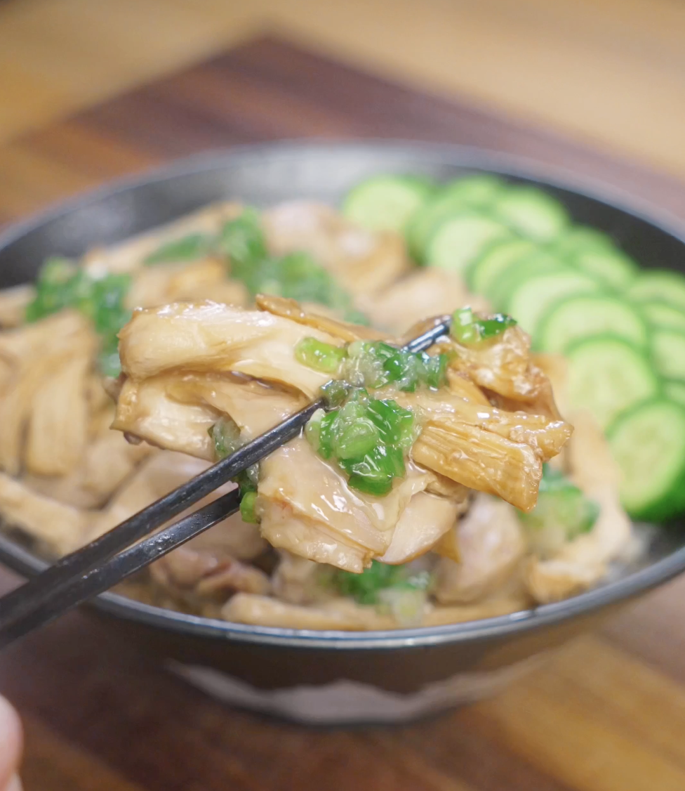 Close-up bite of tender poached ginger scallion chicken lifted with chopsticks, topped with glossy ginger scallion oil and served over steamed rice with sliced cucumbers in the background.
