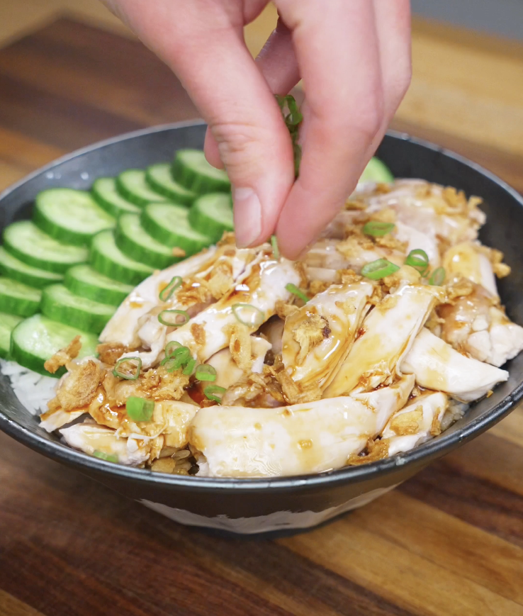 Finished bowl of golden oil poached chicken topped with crispy caramelized aromatics and fresh scallions, served over rice with sliced cucumbers for a comforting high-protein Asian meal.