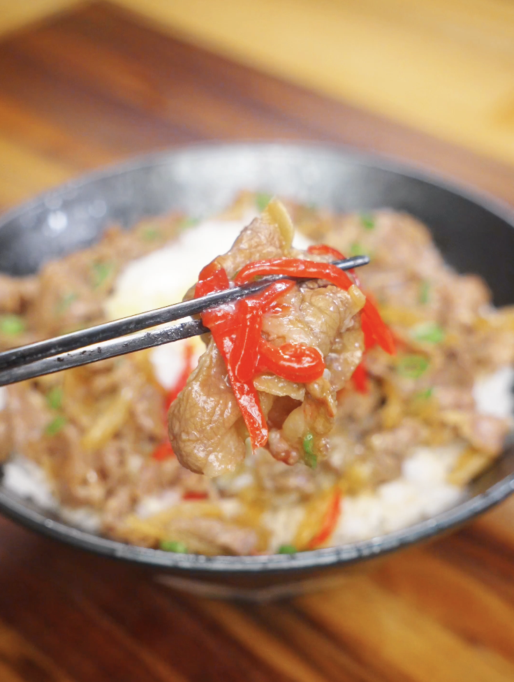 Close-up chopsticks bite of tender gyudon beef and onions coated in a savory soy-based sauce, served over steamed white rice.
