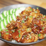Finished bowl of honey garlic chicken served over white rice with sliced cucumbers, sesame seeds, and scallions, showcasing a glossy caramelized honey garlic glaze as a healthy Asian dinner.