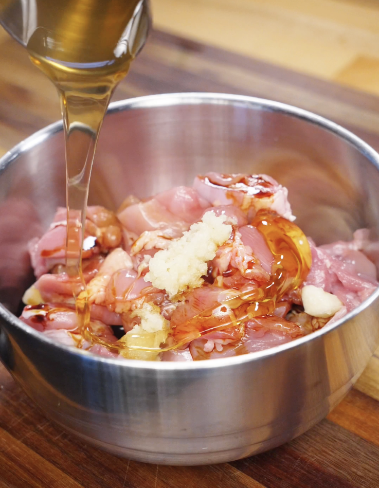 Raw cubed chicken thighs in a stainless steel bowl being marinated with honey, soy sauce, minced garlic, and vinegar as the base for a sticky honey garlic chicken sauce.