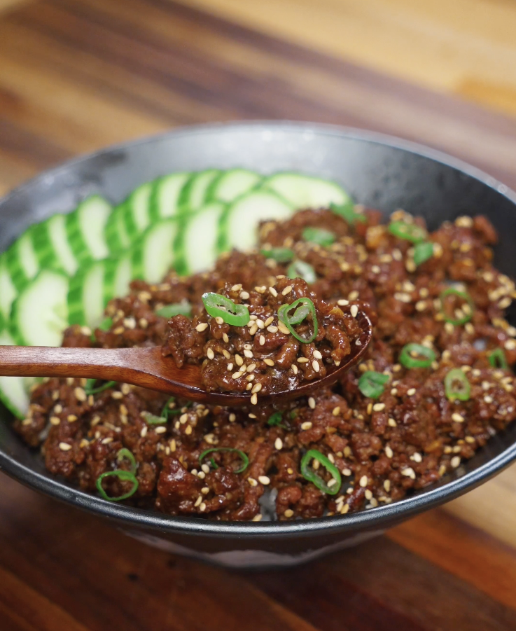 Close-up spoonful of Korean BBQ beef showing caramelized ground beef coated in a rich soy-sesame sauce, garnished with sesame seeds and scallions over a bowl of rice.