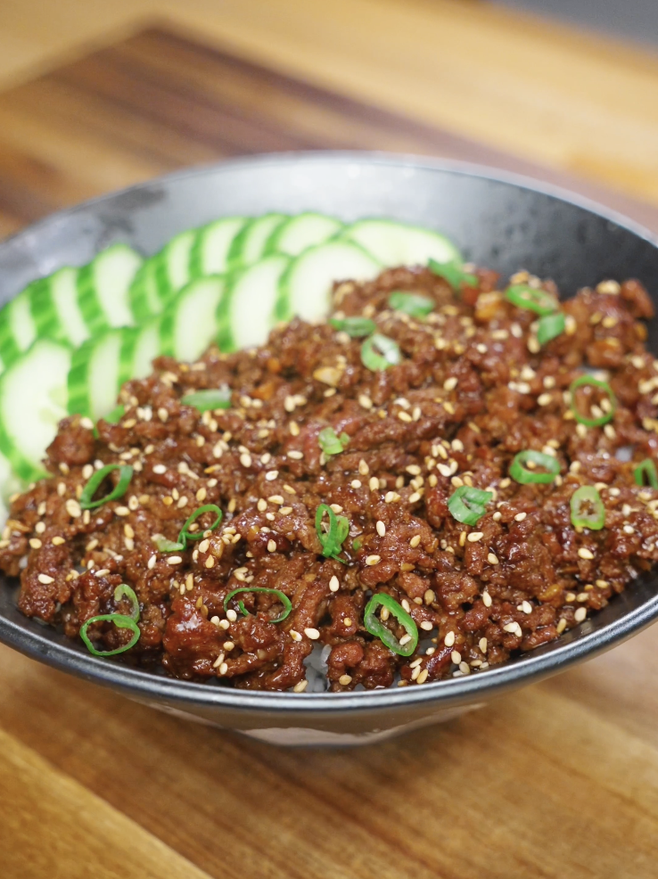 Finished bowl of Korean BBQ beef made with savory, slightly sweet ground beef cooked in a glossy soy-sesame sauce, served over steamed rice with sliced cucumbers, scallions, and sesame seeds as a healthy Asian dinner.
