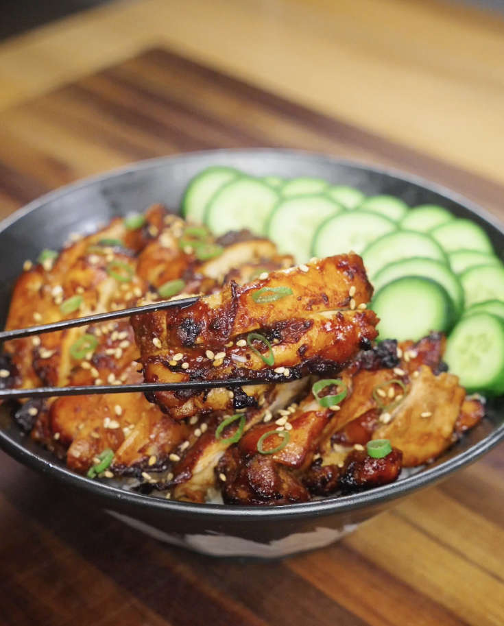 Close-up chopsticks bite of juicy Korean BBQ chicken showing caramelized edges, sticky gochujang glaze, and tender chicken thigh over rice.