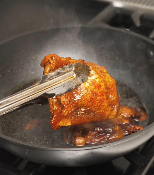 A soy sauce braised chicken thigh being lifted from the pan with tongs, showing glossy skin and rich color after simmering in the soy-forward sauce.
