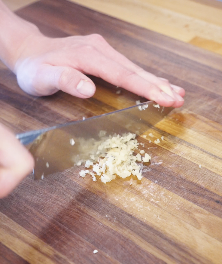 Finely minced garlic and ginger being chopped on a wooden cutting board to build the aromatic base for Korean BBQ beef.