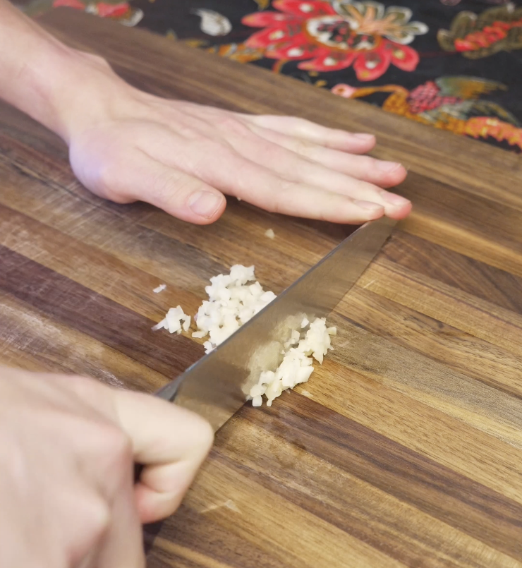 Finely mincing fresh garlic on a wooden cutting board to build flavor for a classic string bean chicken stir fry.