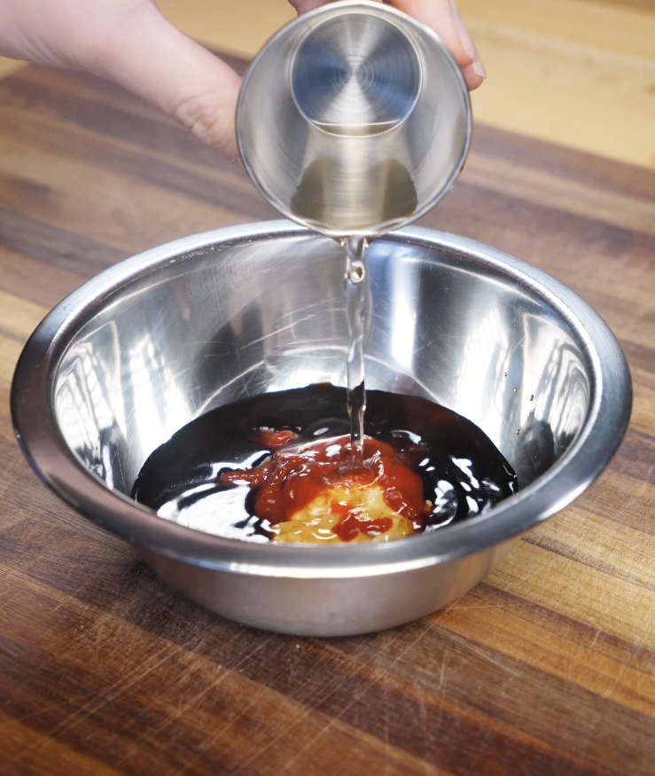 Peanut butter, gochujang, soy sauces, vinegar, sugar, and water being poured into a stainless steel bowl to make a creamy spicy peanut sauce for Spicy Peanut Chicken.