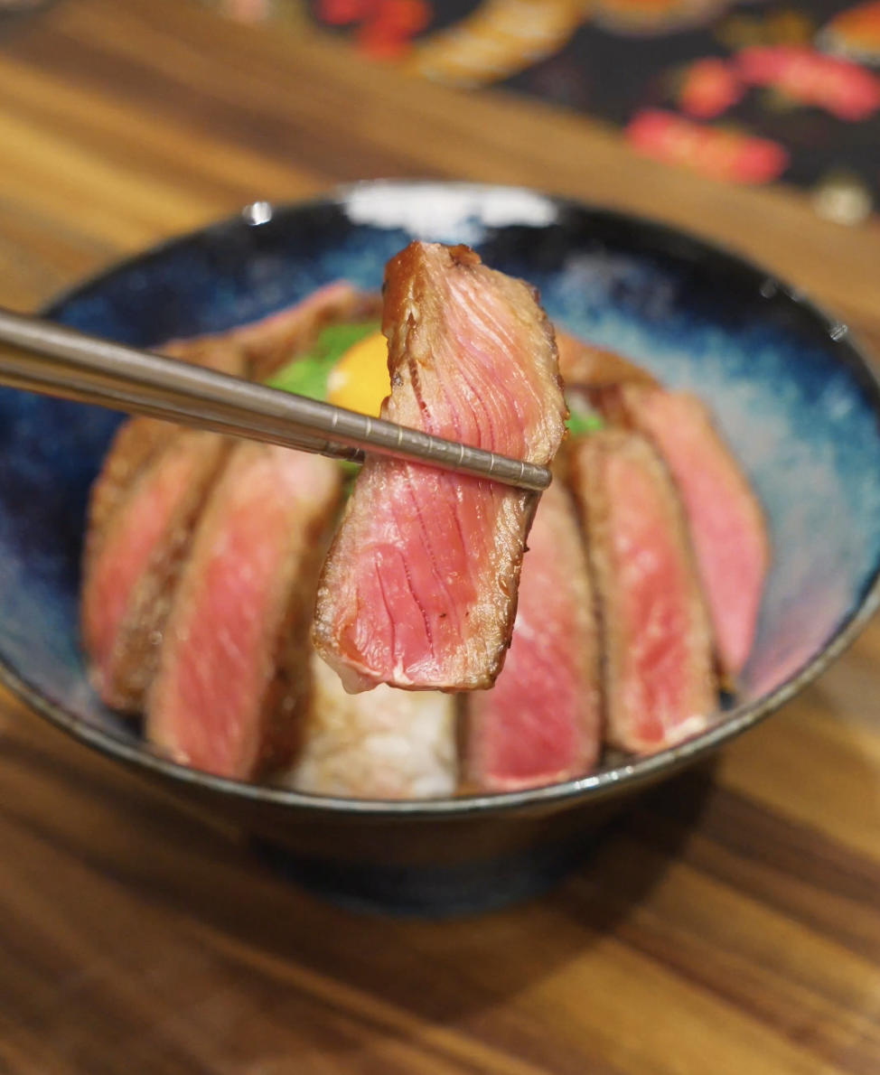 A slice of medium-rare steak held with chopsticks above the rice bowl, showing a tender pink center and seared exterior.