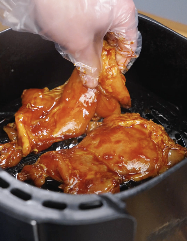 Gloved hand placing marinated Korean BBQ chicken thighs into an air fryer basket before cooking until caramelized and tender.