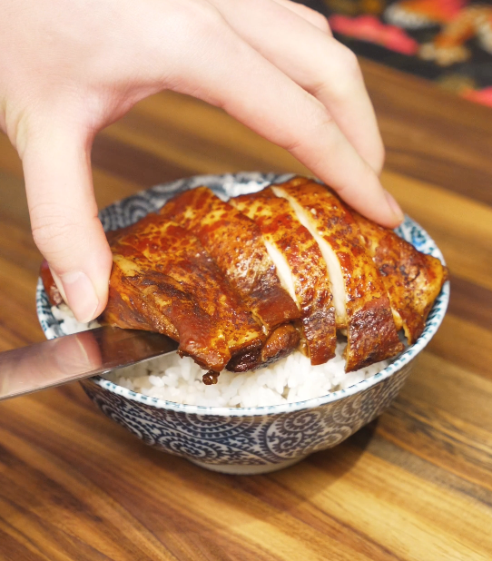 Sliced soy sauce chicken being placed over a bowl of steamed white rice, ready to serve with optional greens.