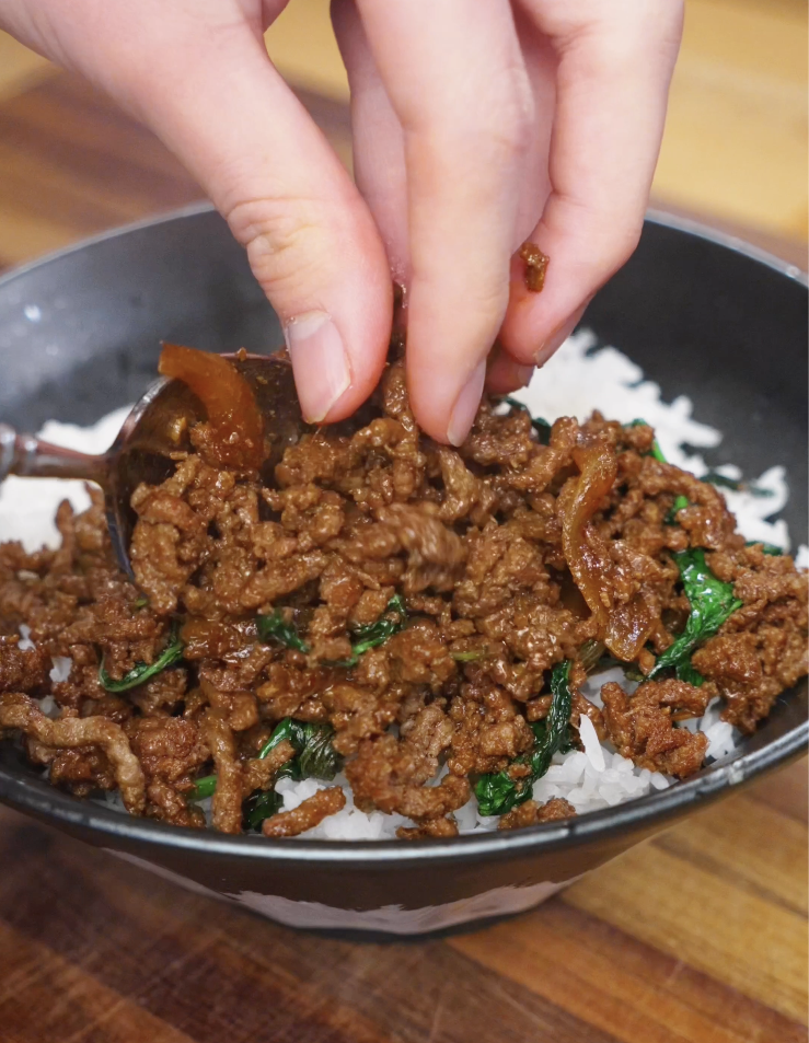 Pad Kra Pao beef being spooned over steamed white rice, preparing the dish for serving as a high-protein Thai dinner.