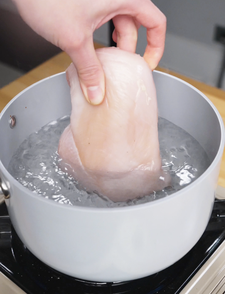 Chicken breast being gently lowered into a pot of boiling salted water to poach until tender and fully cooked for Vietnamese chicken salad.