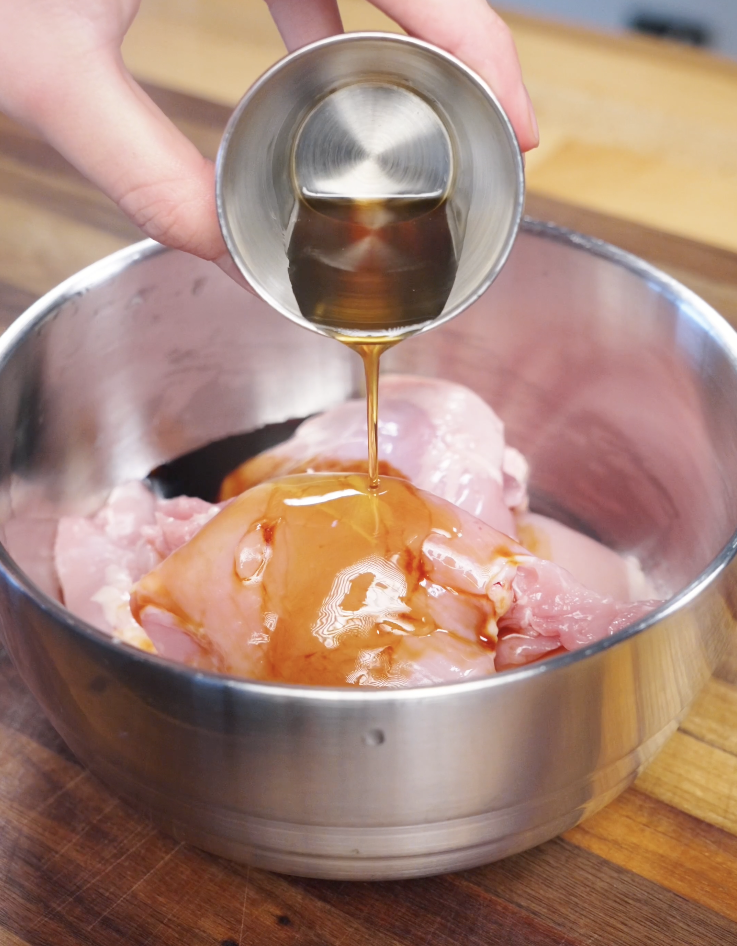 Dark soy sauce, light soy sauce, gochujang, and sesame oil being poured over raw chicken thighs in a stainless steel bowl to marinate Korean BBQ chicken.