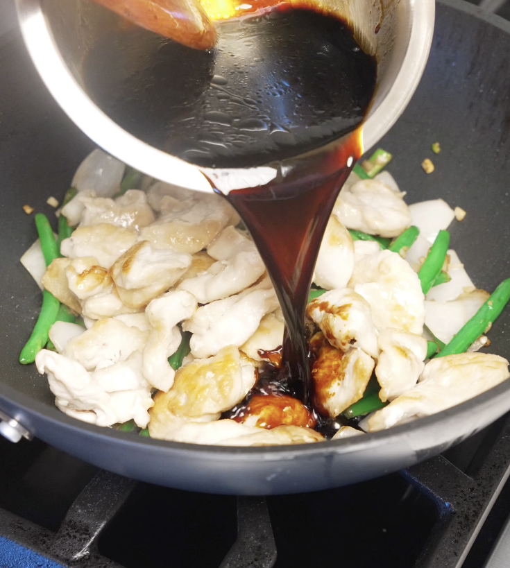 Dark soy-based stir fry sauce being poured into the pan with chicken, onions, and string beans to coat everything evenly.