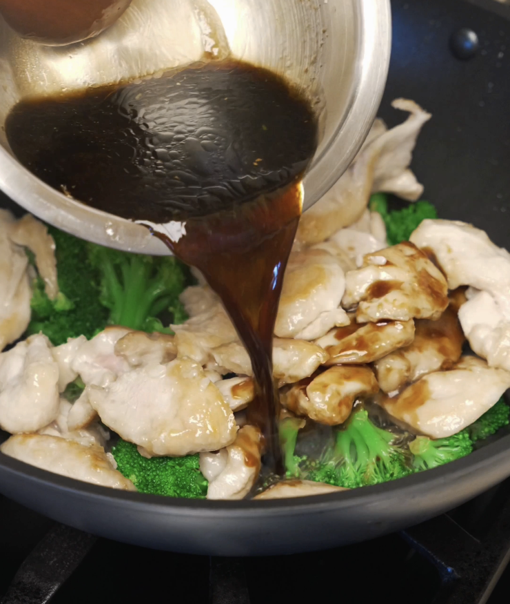 Soy-based chicken and broccoli sauce being poured into a pan with partially cooked chicken and bright green broccoli florets.