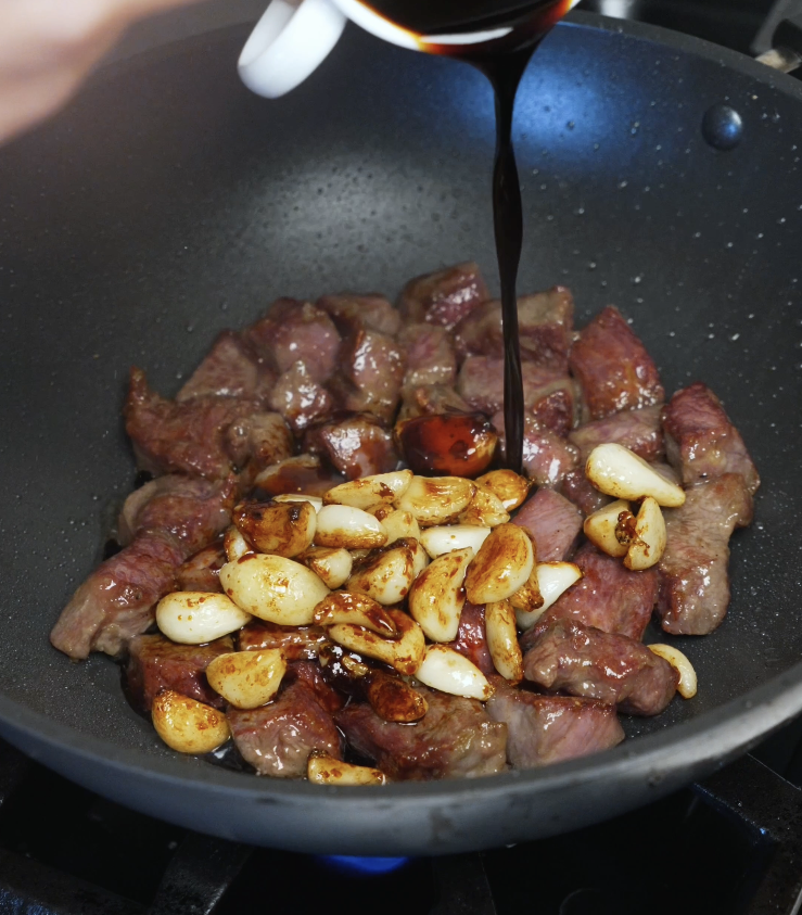 Dark savory stir fry sauce being poured over seared steak cubes and caramelized garlic in a hot pan.