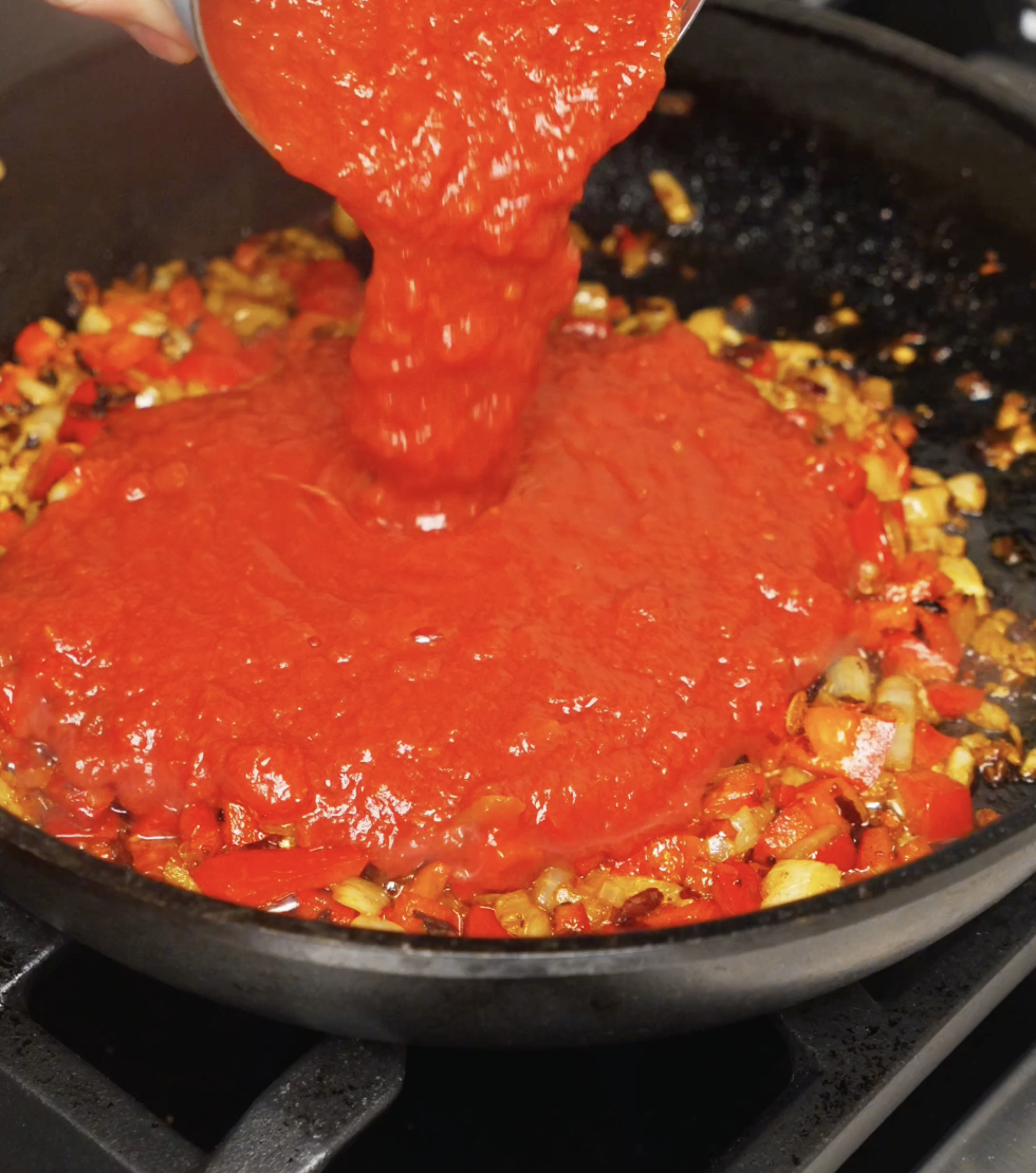 Crushed tomatoes being poured into a pan of saut&eacute;ed onions, red bell peppers, garlic, and spices, forming the base for chili oil shakshuka.