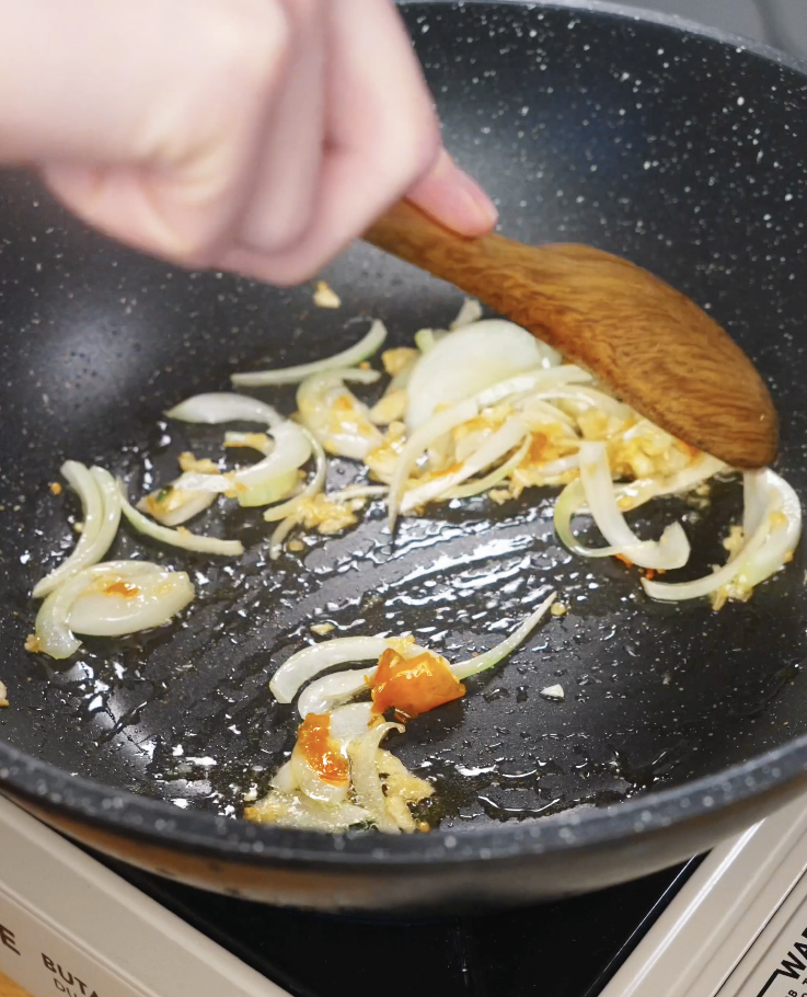 Sliced onions and chili-garlic paste being saut&eacute;ed in oil in a pan until soft and fragrant, forming the aromatic base of Pad Kra Pao.