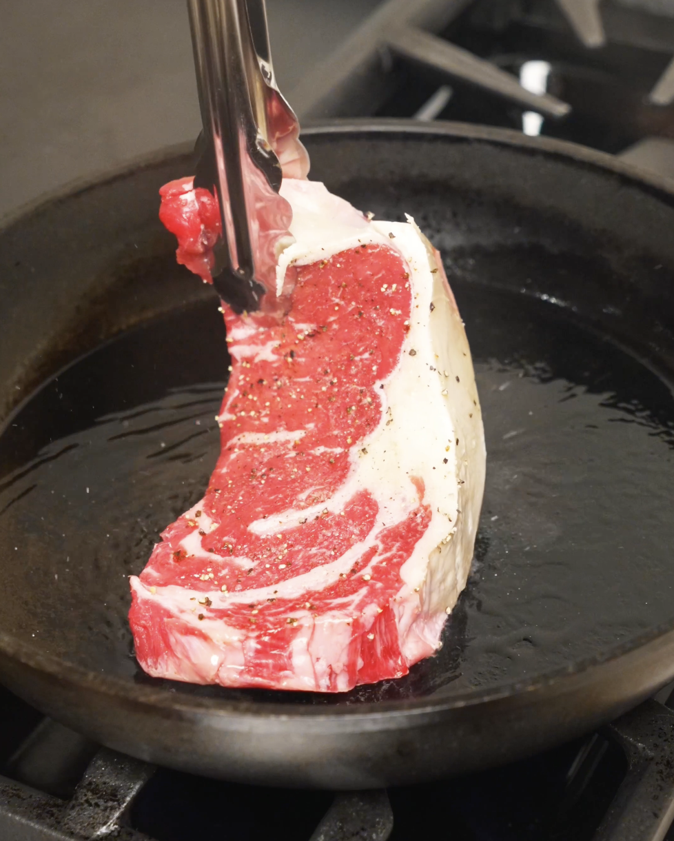 NY strip steak held with tongs and placed into a hot pan with oil, beginning the searing process to build a flavorful crust.
