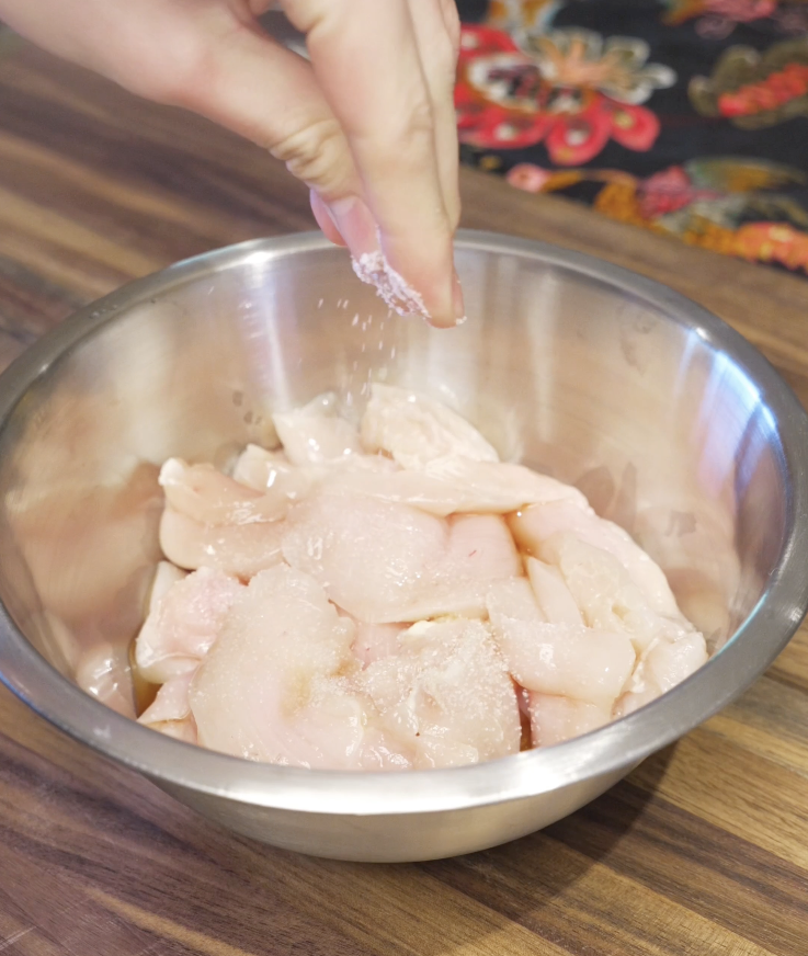 Thinly sliced chicken breast being seasoned with salt in a stainless steel bowl as the first step in making string bean chicken stir fry.