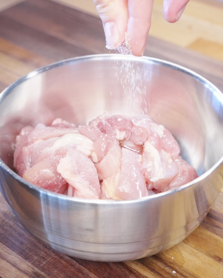 Cubed boneless chicken thighs being seasoned with salt in a stainless steel bowl as the first step in making garlic chicken stir fry.