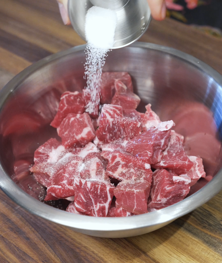 Salt being sprinkled over raw cubed flatiron steak in a stainless steel bowl before marinating.