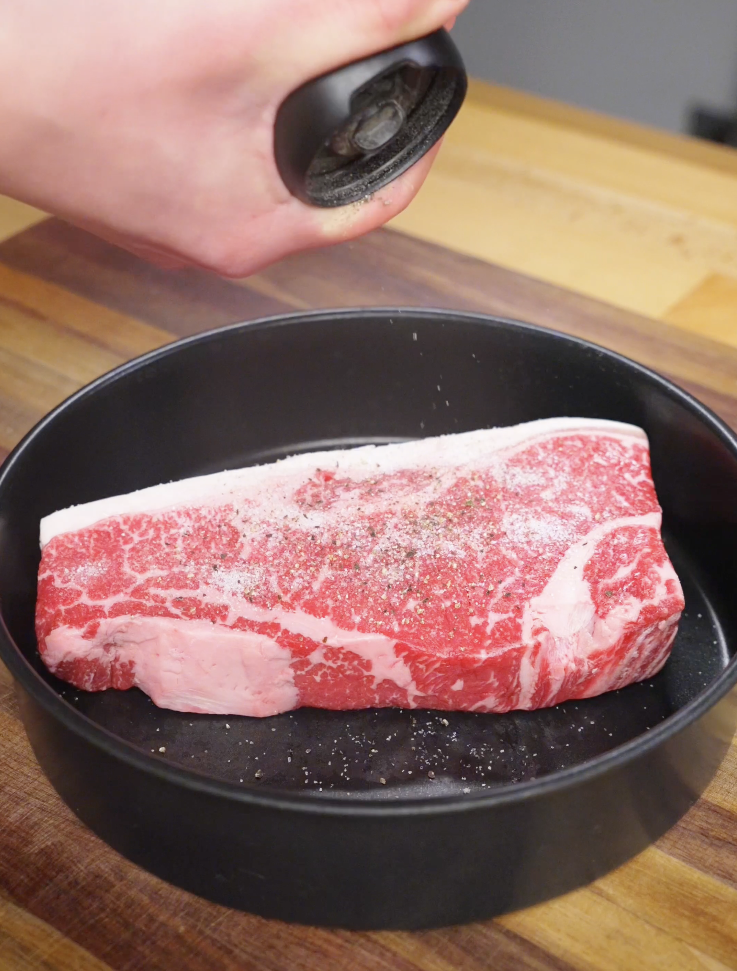 Raw sirloin steak being seasoned generously with salt and black pepper before cooking for Crying Tiger Steak.