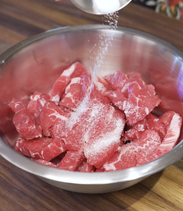 Thinly sliced flank steak being seasoned with salt and cornstarch in a metal mixing bowl to prepare tender beef for beef and broccoli stir fry.