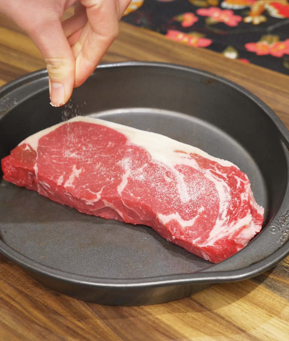 Raw NY strip steak being seasoned with salt and black pepper in a pan before searing for the Japanese steak bowl.