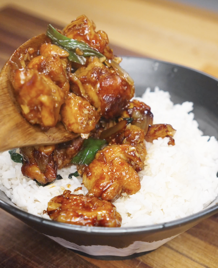 Spoonful of garlic chicken stir fry being served over steamed white rice, showing glossy sauce-coated chicken and onions.