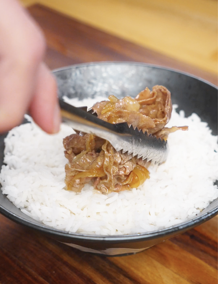Placing tender gyudon beef and onions onto a bowl of freshly steamed white rice using kitchen tongs.