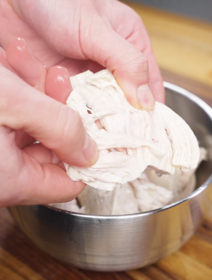 Hands pulling apart a poached chicken breast into thin shreds after cooking for Vietnamese chicken salad.