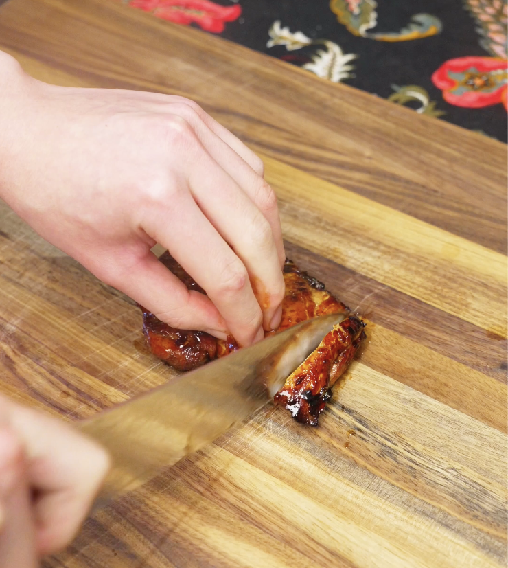 Knife slicing rested lemongrass chicken thighs into thin strips on a wooden cutting board.