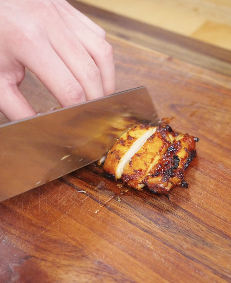 Cooked Korean BBQ chicken thigh being sliced on a wooden cutting board, showing juicy interior and caramelized exterior.