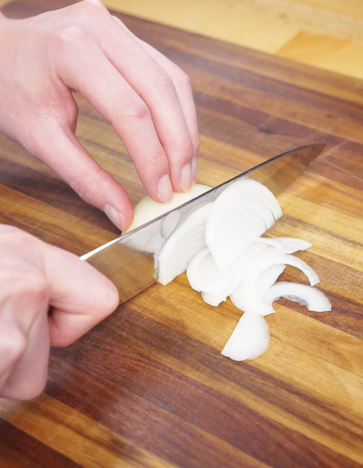 Thinly slicing a white onion on a wooden cutting board as the first prep step for making classic Japanese gyudon beef bowl.
