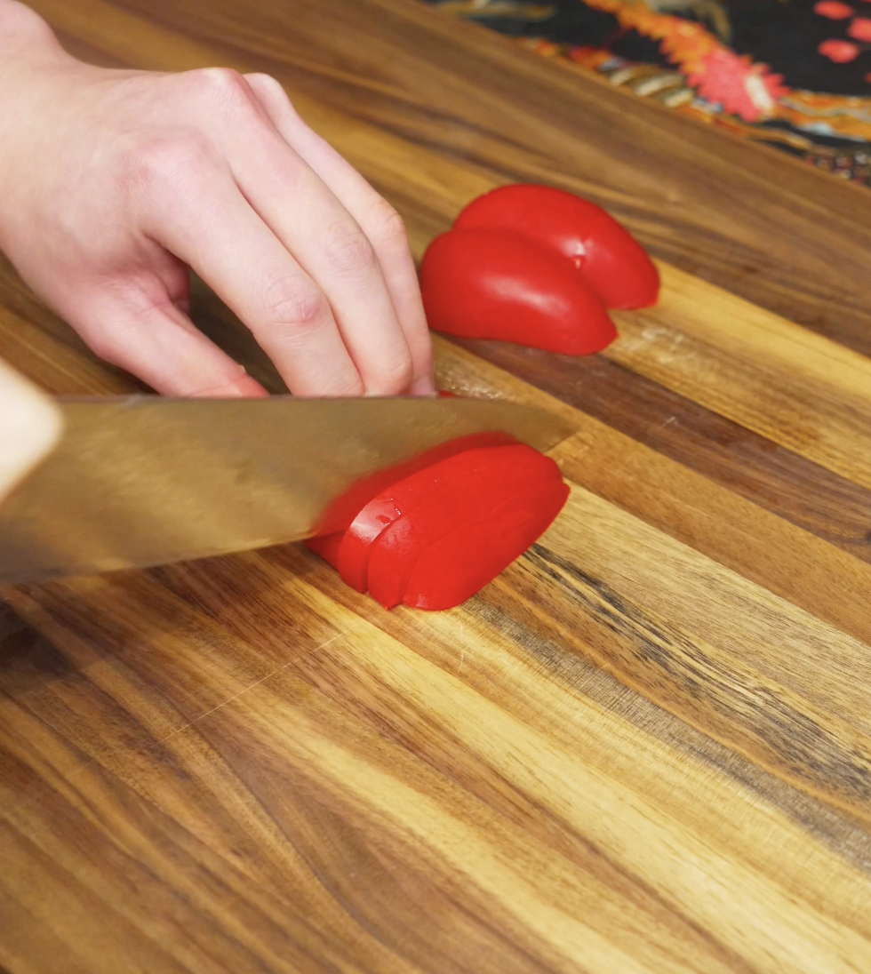 Red bell pepper being sliced on a wooden cutting board as prep for the chili oil shakshuka vegetable base.