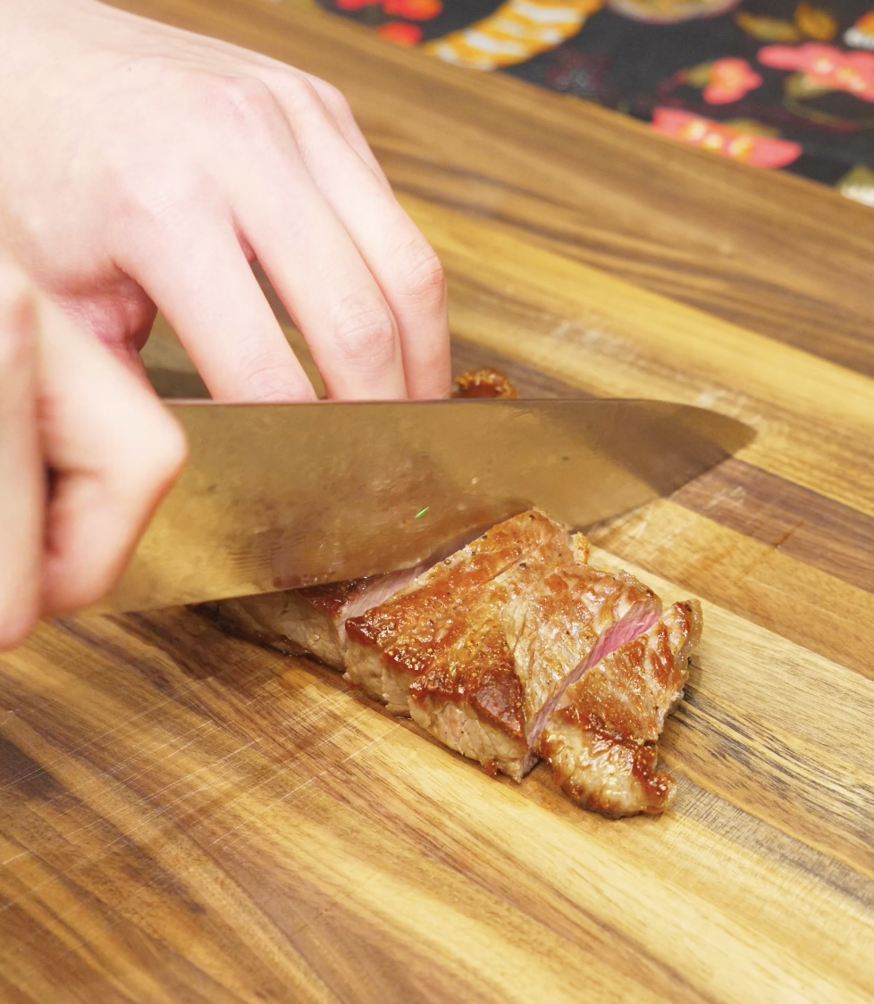 Seared steak being sliced thin against the grain on a wooden cutting board, showing a juicy medium-rare center.