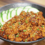 Finished bowl of Spicy Peanut Chicken made with ground chicken cooked in a creamy gochujang peanut sauce, served over steamed rice with sliced cucumbers, sesame seeds, and scallions as a healthy Asian dinner.