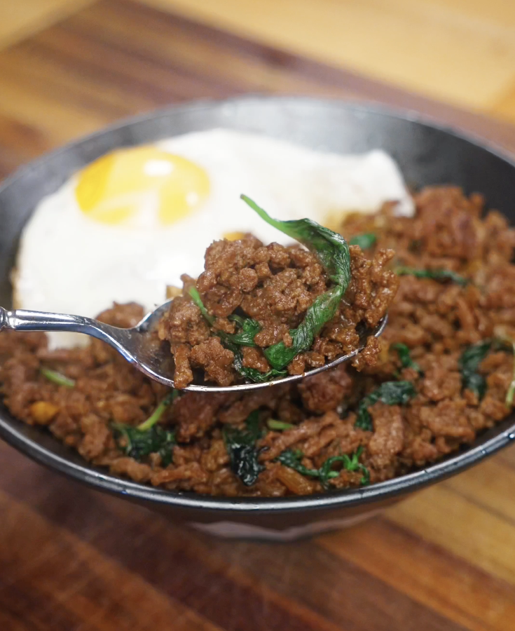 Spoon lifting a bite of savory ground beef Pad Kra Pao mixed with wilted basil leaves, highlighting the juicy texture and bold seasoning.