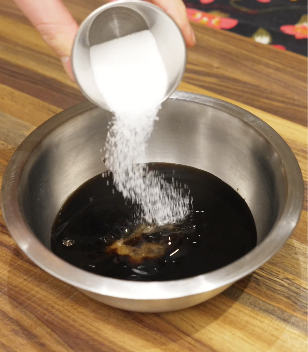 Granulated sugar being poured into a bowl of soy sauce, mirin, and sake to create the lightly sweet glaze for the Japanese steak bowl.