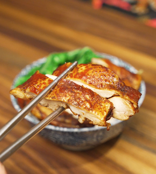 A slice of soy sauce chicken lifted with chopsticks, showing juicy meat and glossy soy glaze over rice with steamed greens in the background.