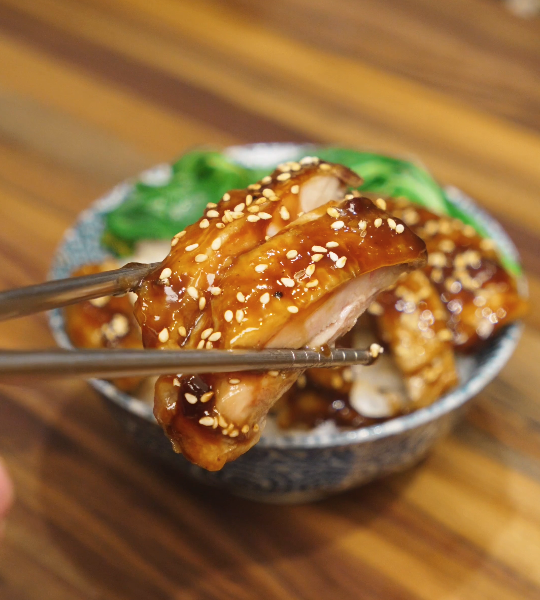 A slice of teriyaki chicken thigh lifted with chopsticks, showing juicy meat coated in a glossy homemade teriyaki sauce and finished with sesame seeds.