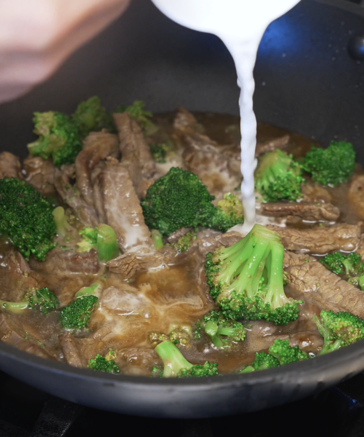 Cornstarch slurry being poured into beef and broccoli to thicken the sauce until glossy and evenly coated.