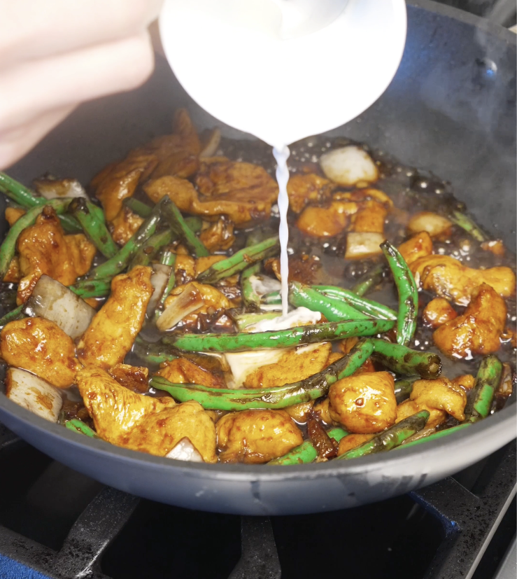 Cornstarch slurry being poured into the pan as the sauce thickens and turns glossy, coating the chicken and vegetables evenly.