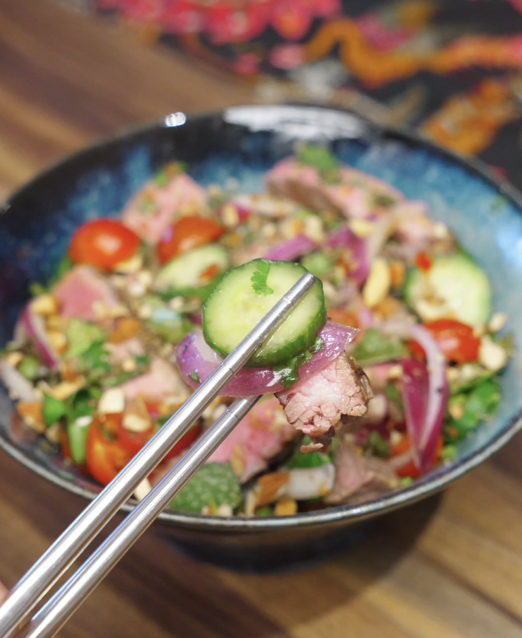 Picking up a bite of Thai beef salad with chopsticks, showing sliced medium-rare steak, cucumber, red onion, herbs, and chopped nuts coated in tangy lime dressing.