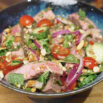 Finished Thai beef salad served in a bowl with sliced steak, red onion, cucumber, cherry tomatoes, fresh herbs, and chopped nuts tossed in a lime-forward dressing.